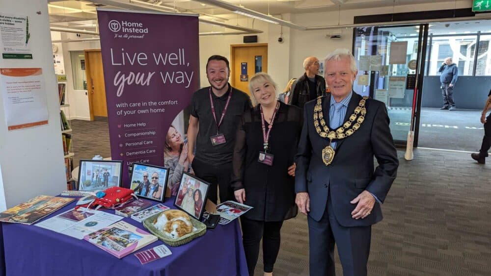 Three people stand by a "Home Instead" care booth with photos, brochures, and a plate of pastries on display. - Home Instead