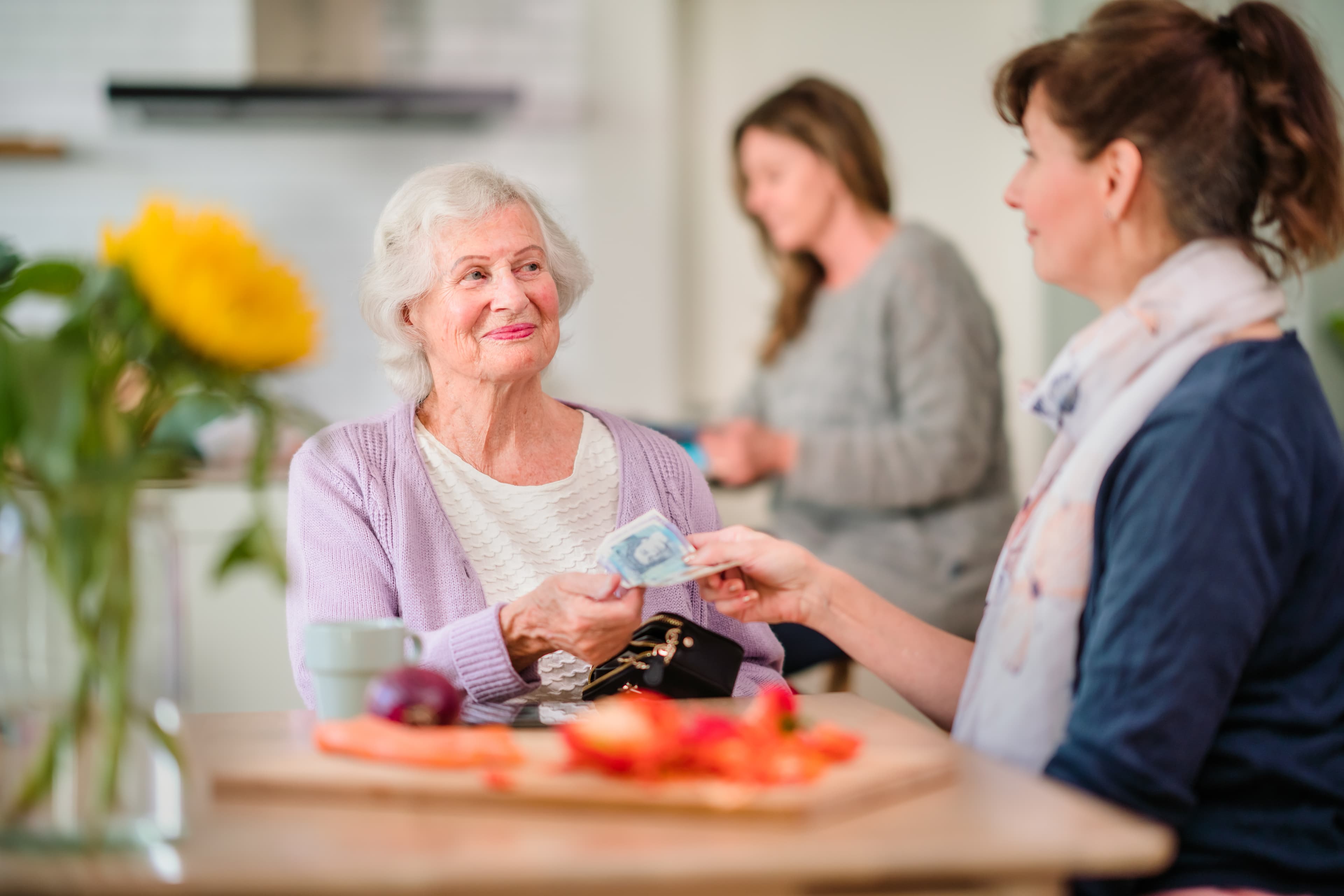 Smiling elderly woman hands cash to another woman at a kitchen table, with a third woman in the background. - Home Instead