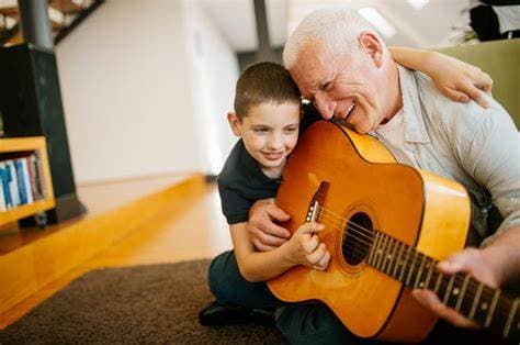 Smiling grandfather and grandson sit on the floor, playing an acoustic guitar together in a cozy room. - Home Instead