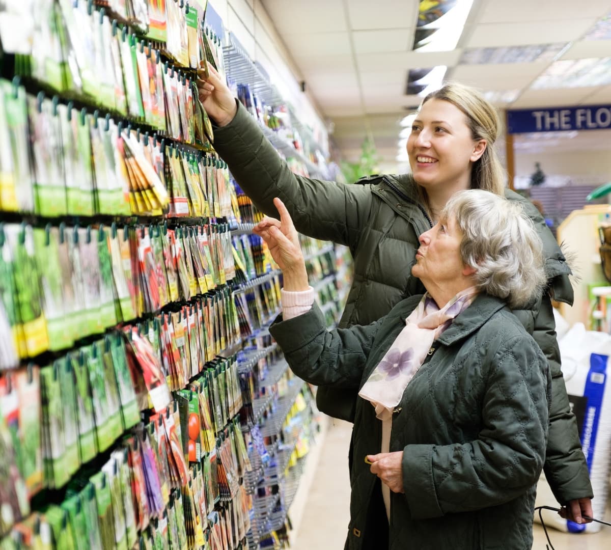 Older woman with short grey hair and wearing winter clothes with her younger female carer with long blonde hair both at the grocery buying seeds