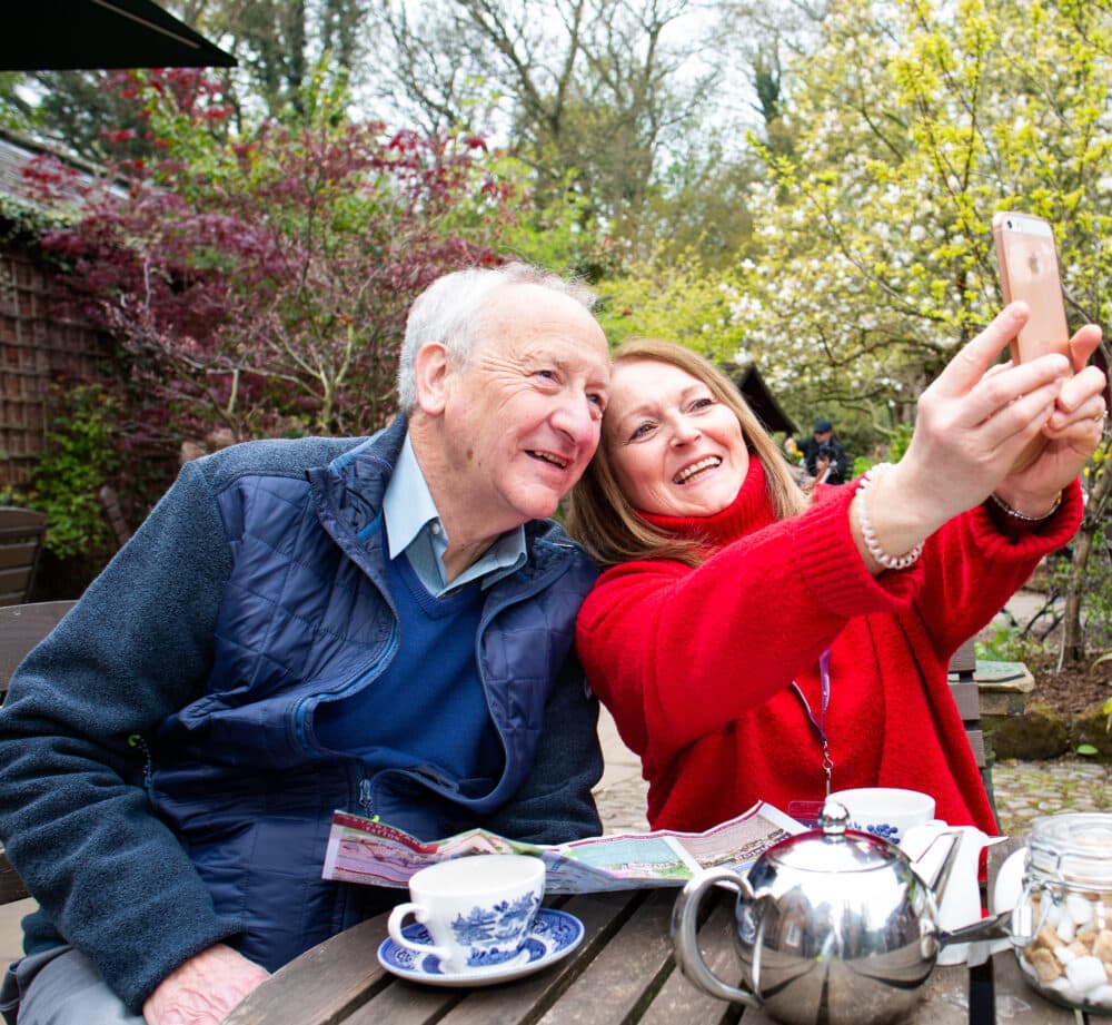Home Instead carer taking a selfie with her client whilst sitting at a picnic table laid out with a tea pot and cups at a garden centre