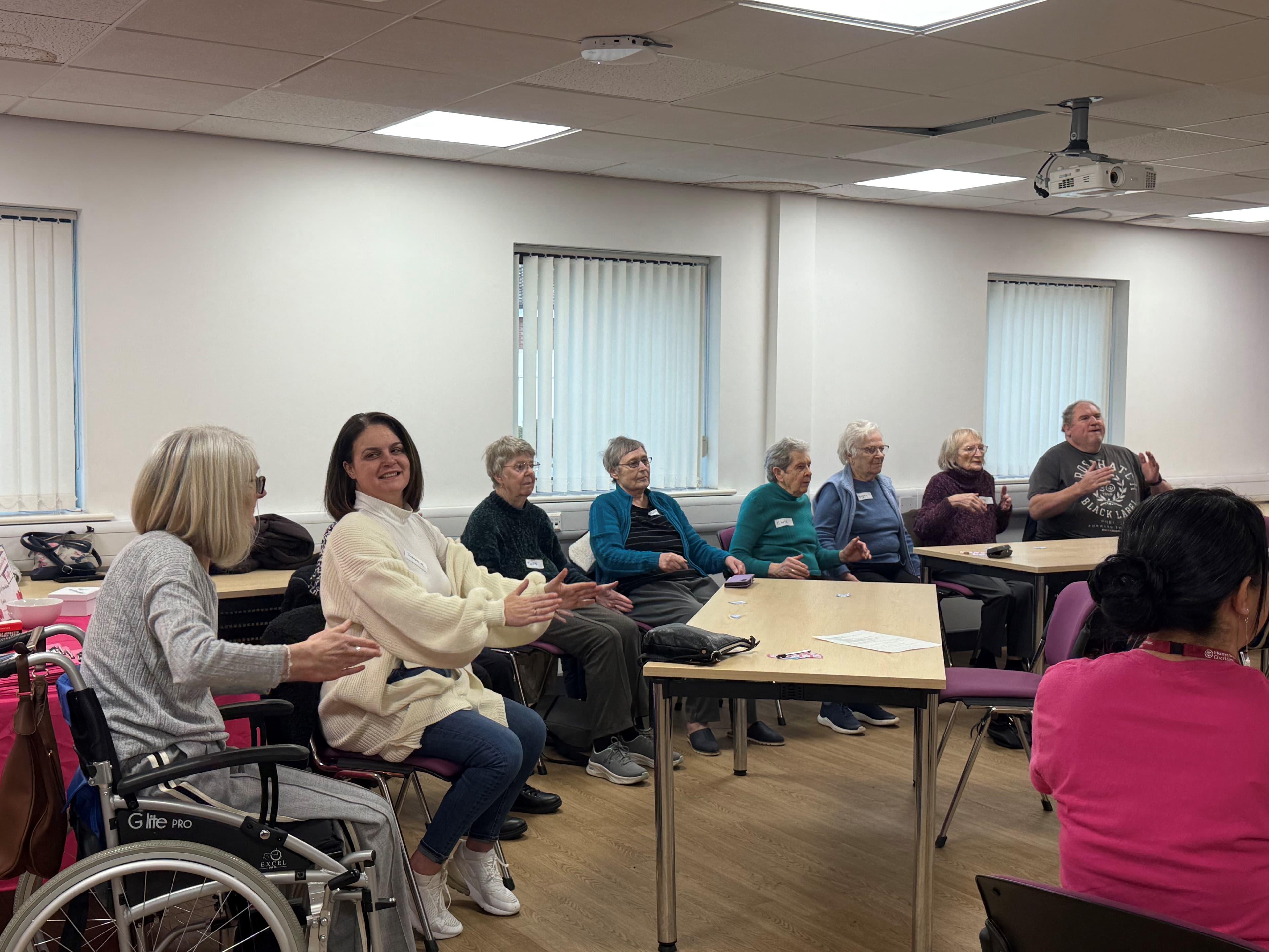 A group of seniors and a younger woman sit in a classroom, smiling and participating in an activity together. - Home Instead