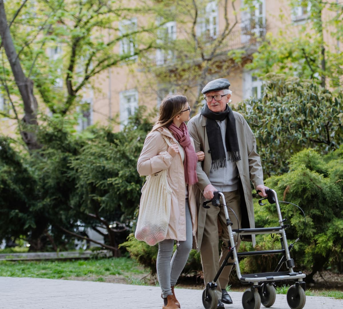 Older man walking outdoors wearing winter clothes and using a walker with the help og his female carer bith smiling and happy