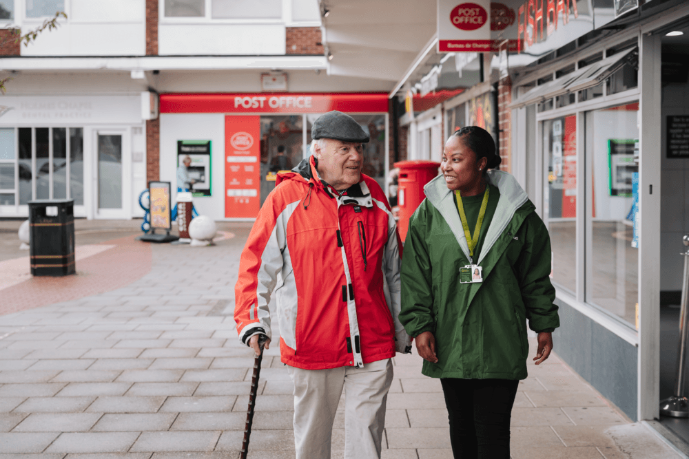 An older man with a cane walks and talks with a carer on a wet pavement outside a post office. - Home Instead