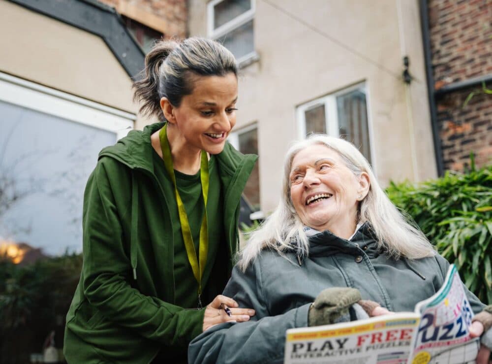 A younger woman smiles at an older woman in a wheelchair who is holding a puzzle magazine outside. - Home Instead