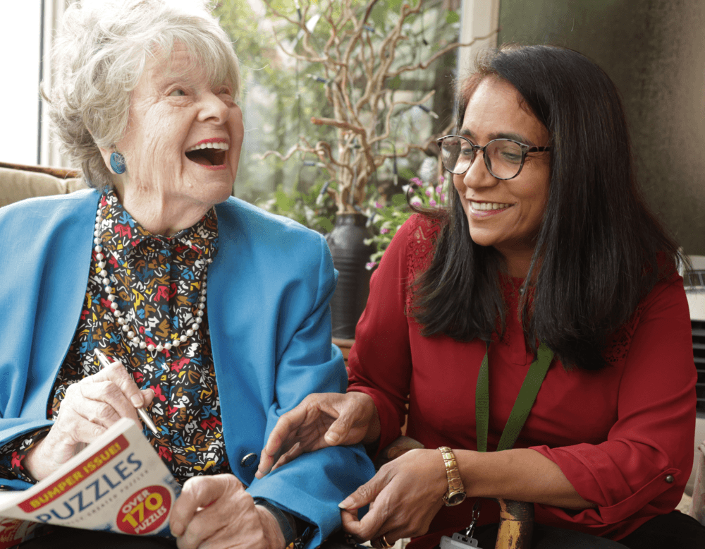 An elderly woman and a younger woman smile and laugh together while holding a puzzle book. - Home Instead