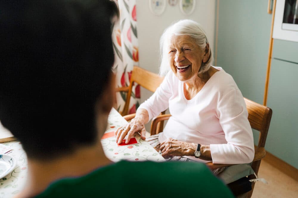 Older woman smiling and talking with another person at a kitchen table decorated with a patterned tablecloth. - Home Instead