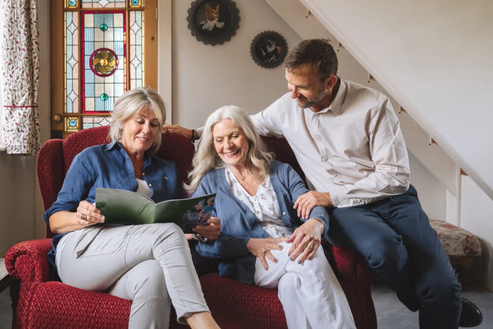 Three adults sit on a red couch, smiling and looking at a book together in a cozy living room. - Home Instead