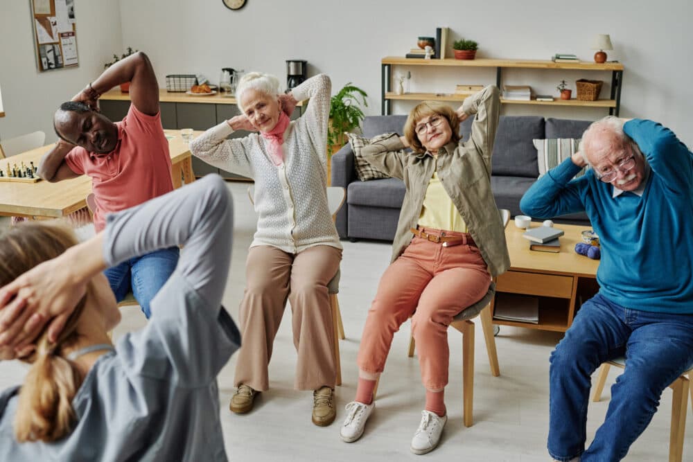 Group of seniors seated indoors, following a leader in stretching exercises with hands behind their heads. - Home Instead