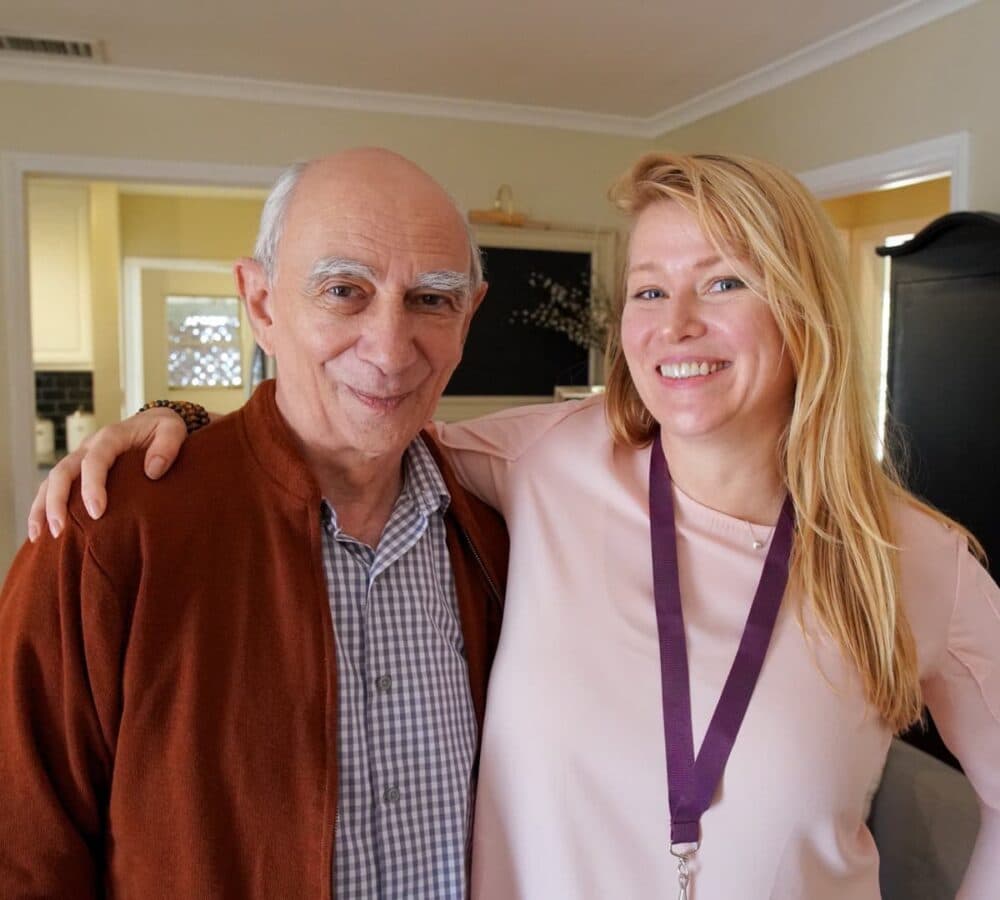 An older man with grey hair and wearing a sweater with his younger female carer wiht long blonde hair wearing pink and both smiling and happy inside the home