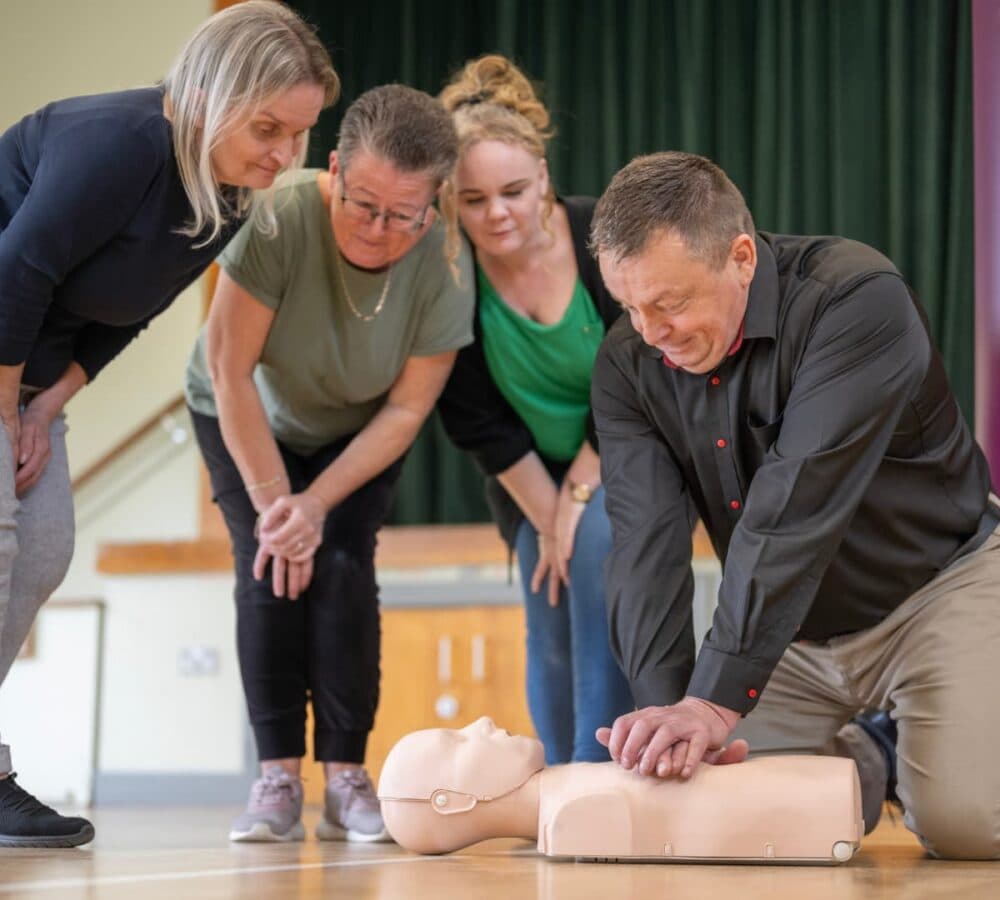 A man demonstrates CPR on a training dummy as three women watch attentively in a classroom setting. - Home Instead