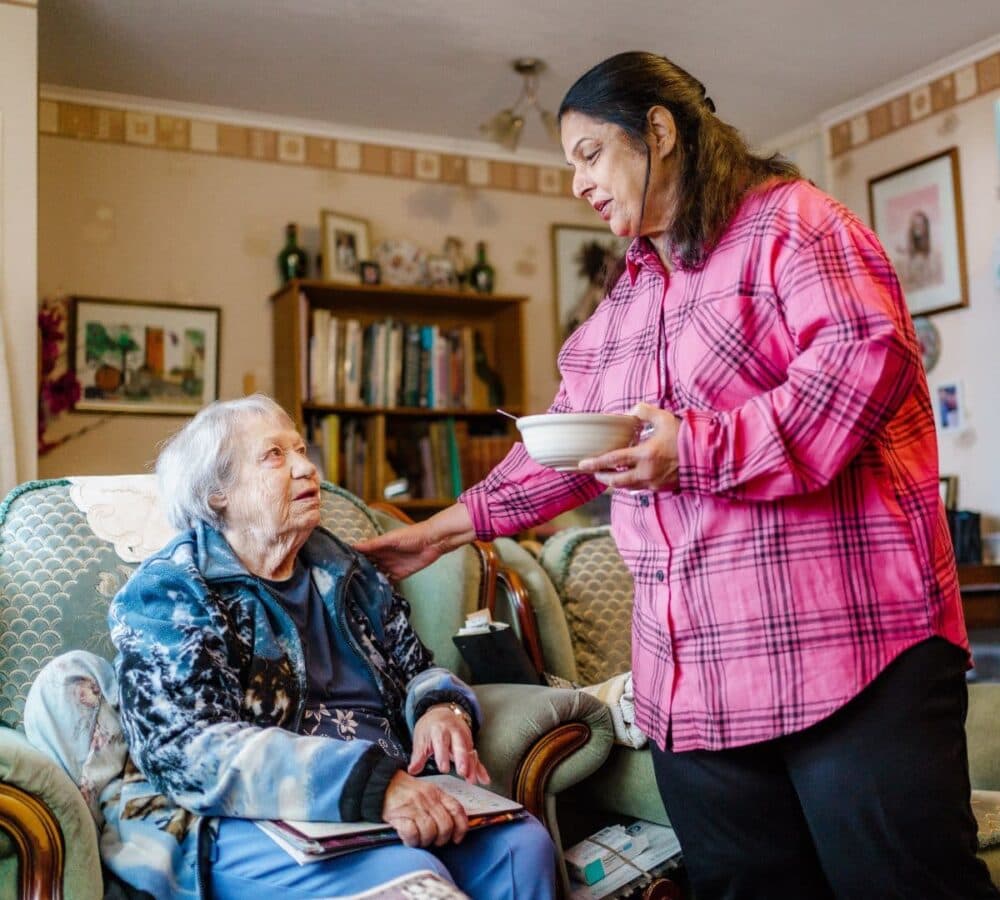 A carer wearing pink long sleeves while holding a cup of coffee for an older female woman sitting on a chair with white hair inside the house