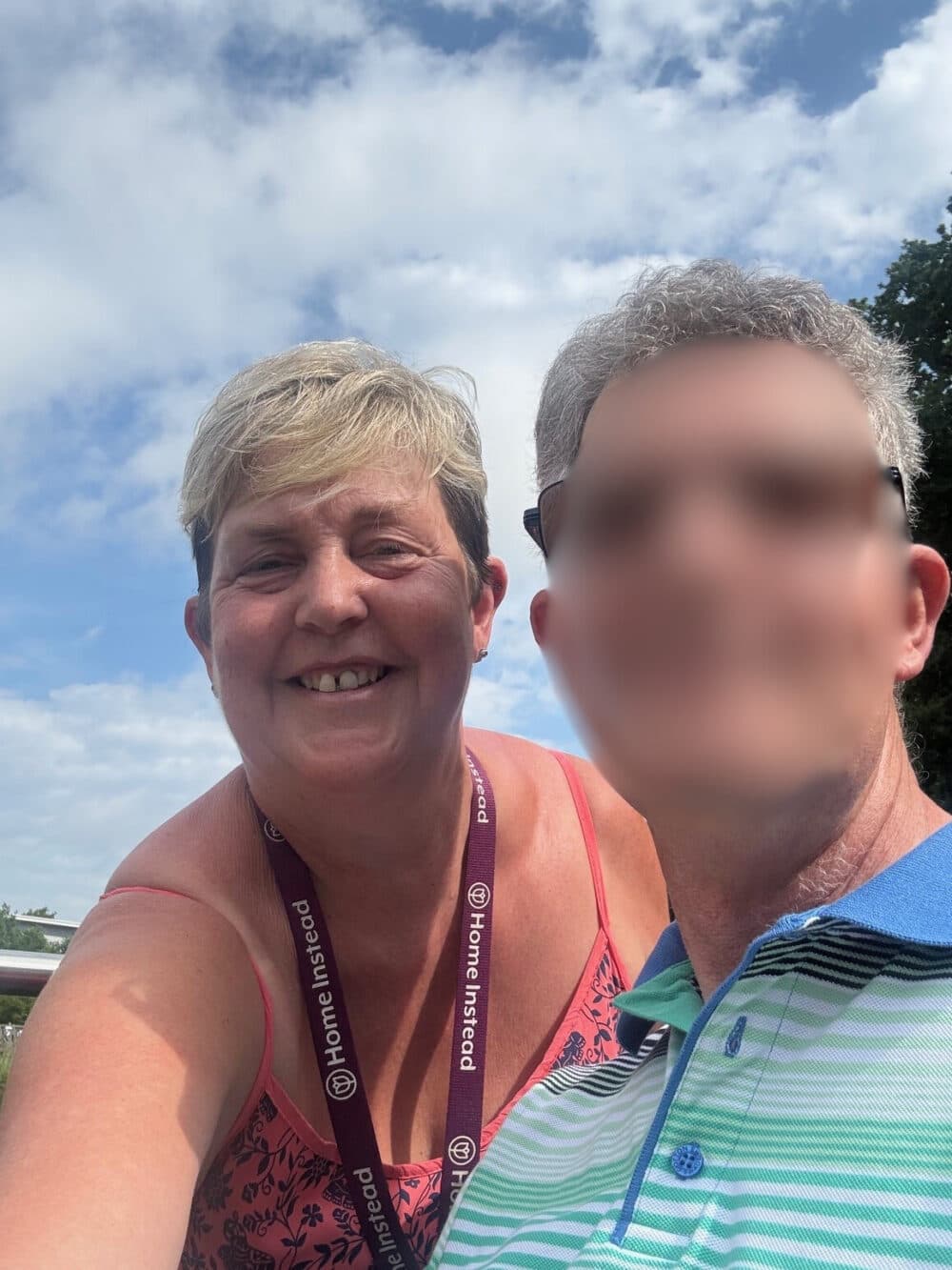 A woman smiling outdoors next to a man with his face blurred, under a partly cloudy sky. - Home Instead