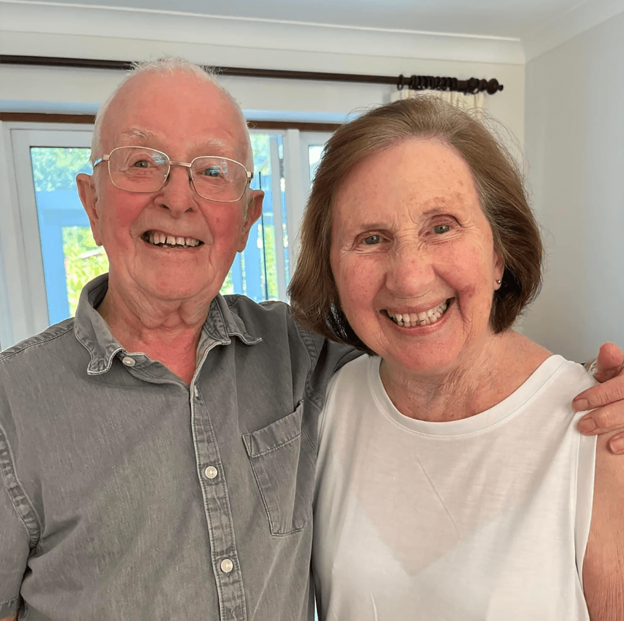 An elderly man and woman smiling together, standing in a living room with a window in the background. - Home Instead