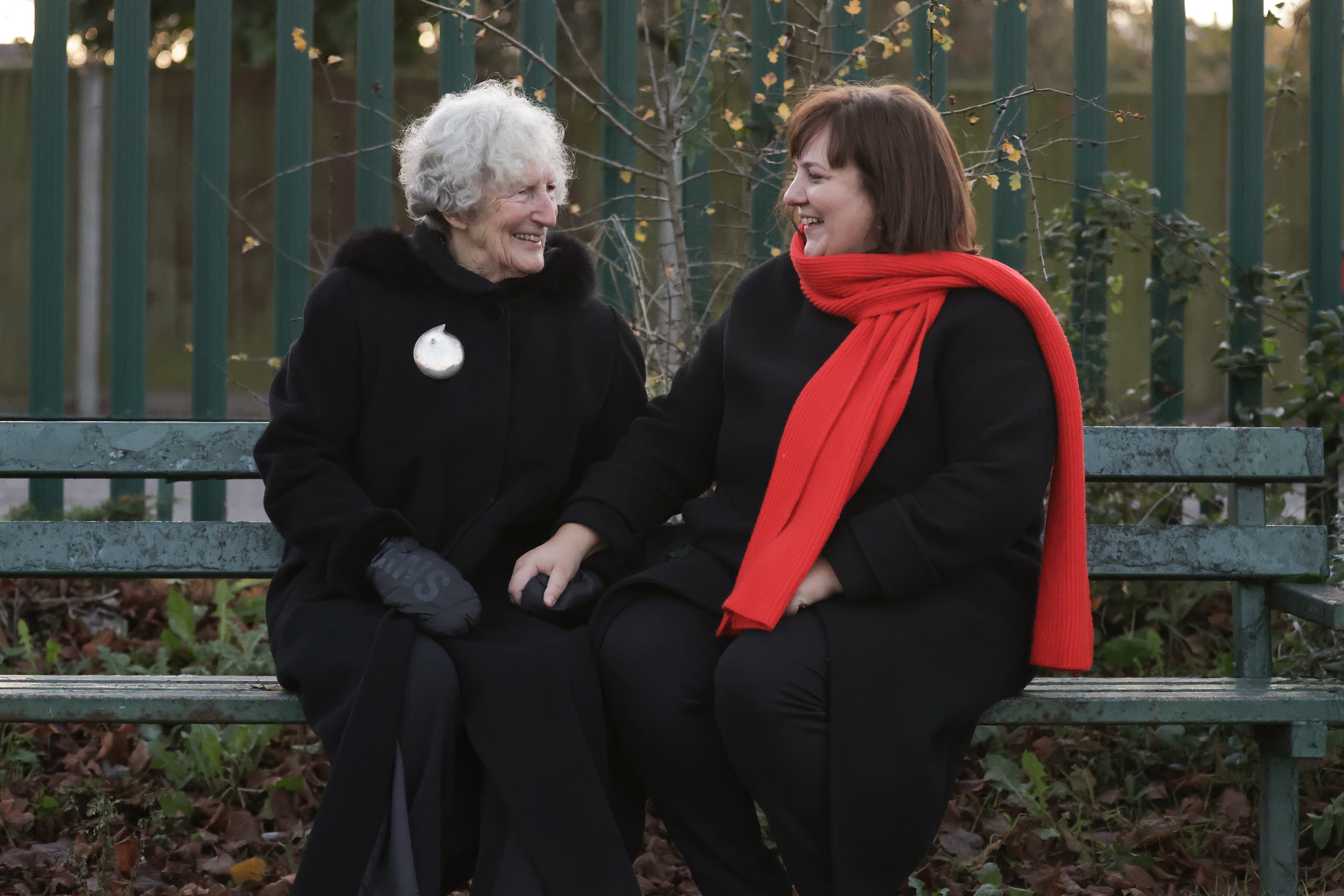 Two women in black coats sit on a bench, smiling, one wearing a red scarf, with greenery in the background. - Home Instead