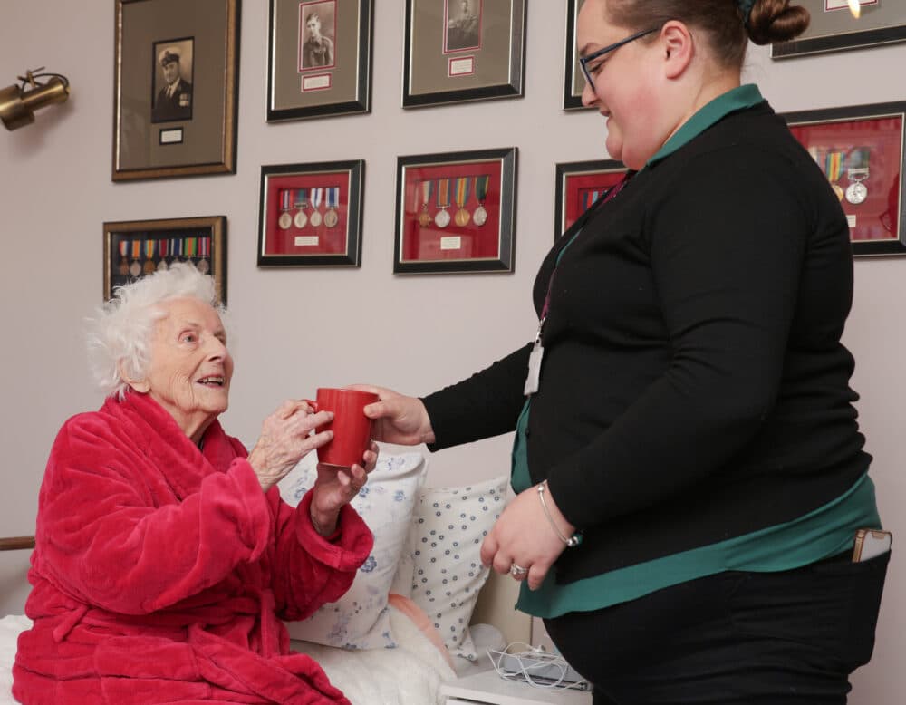 Elderly woman in a red robe receives a mug from a smiling caregiver in a room with framed medals on the wall. - Home Instead