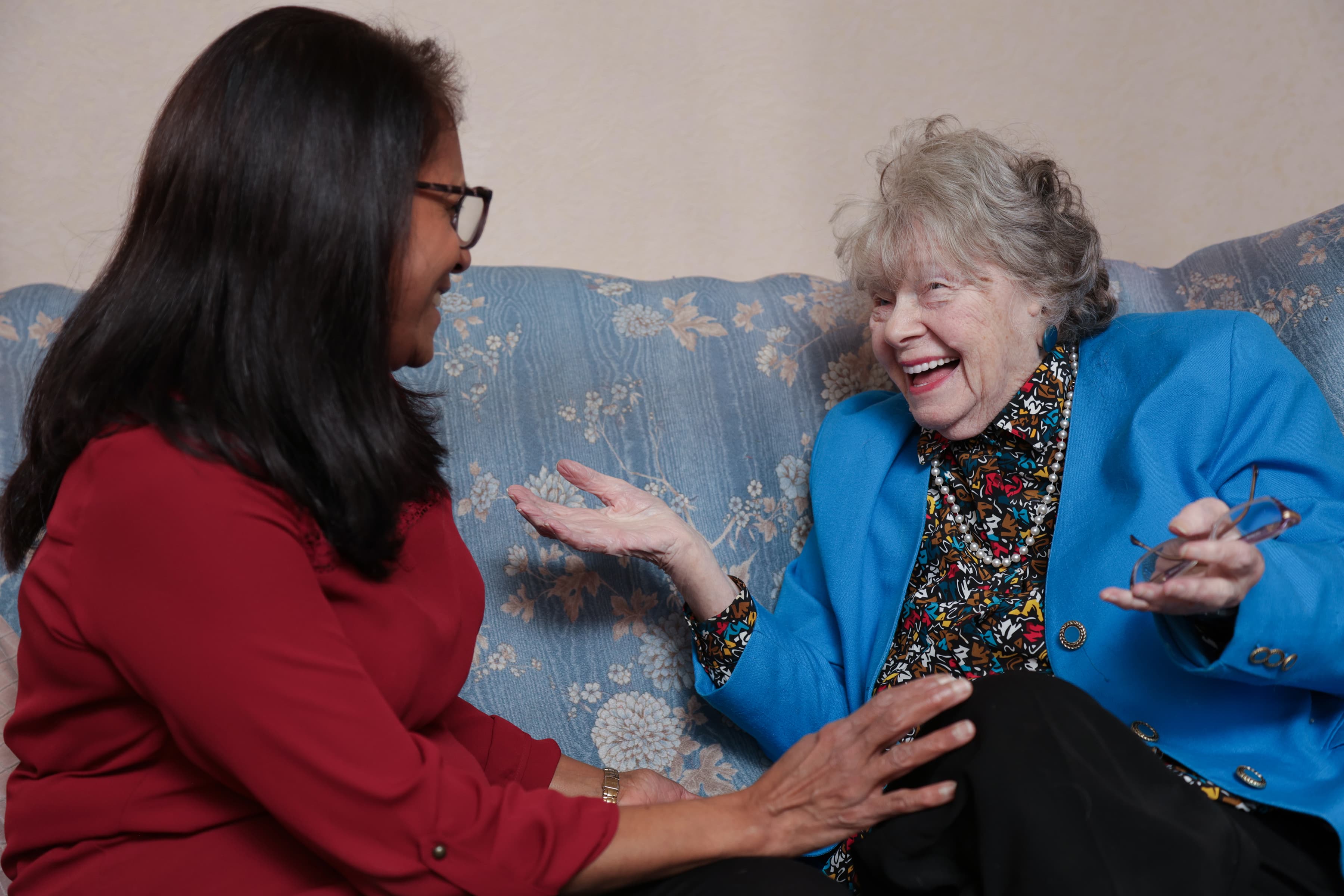 Two women sit on a couch, smiling and chatting warmly, one wearing red and the other in a blue jacket. - Home Instead