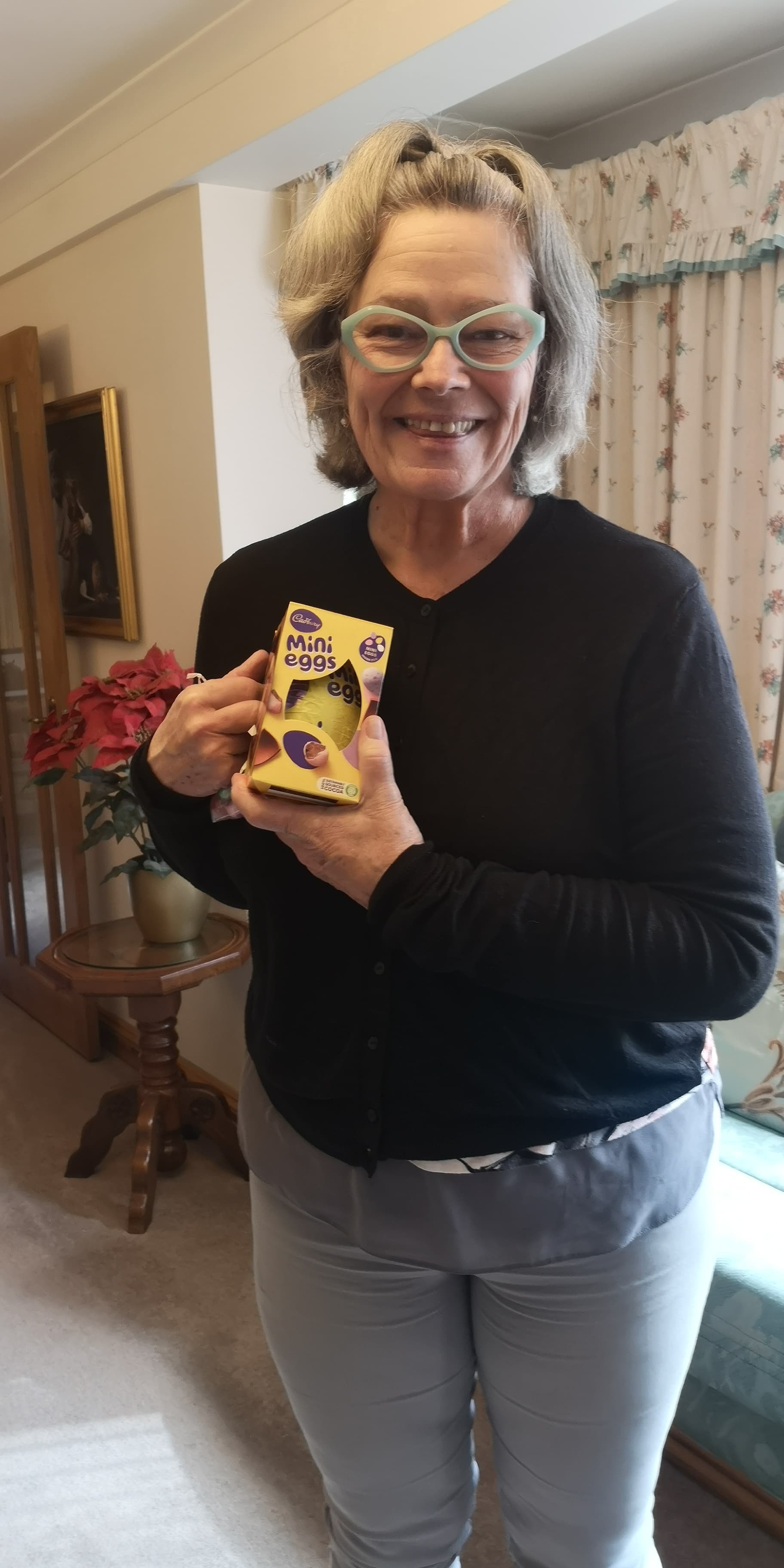 Smiling woman in glasses holding a box of Cadbury Mini Eggs in a cozy living room. - Home Instead