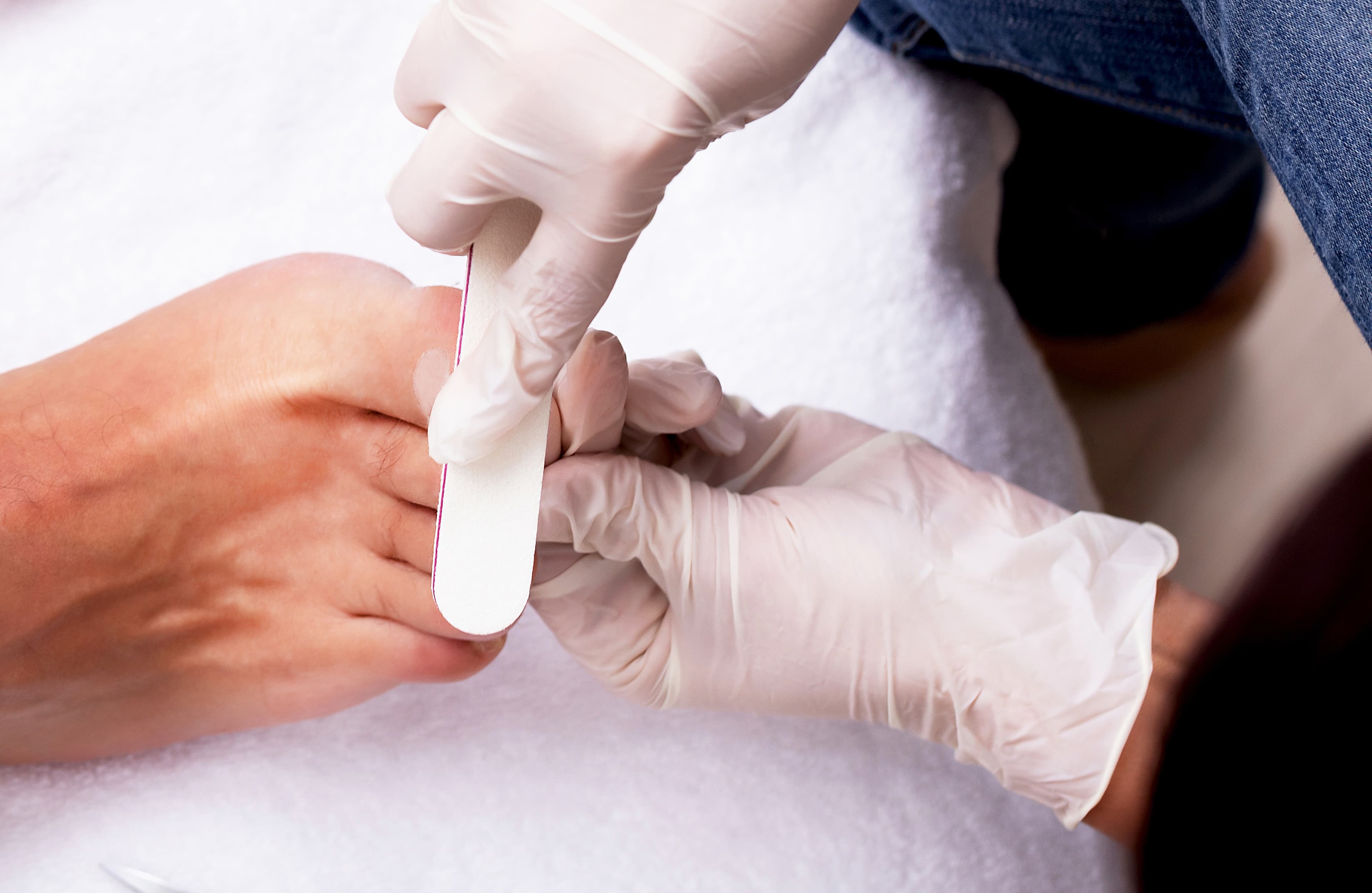 Gloved hands filing toenails during a pedicure on a person's foot, with a white towel underneath. - Home Instead