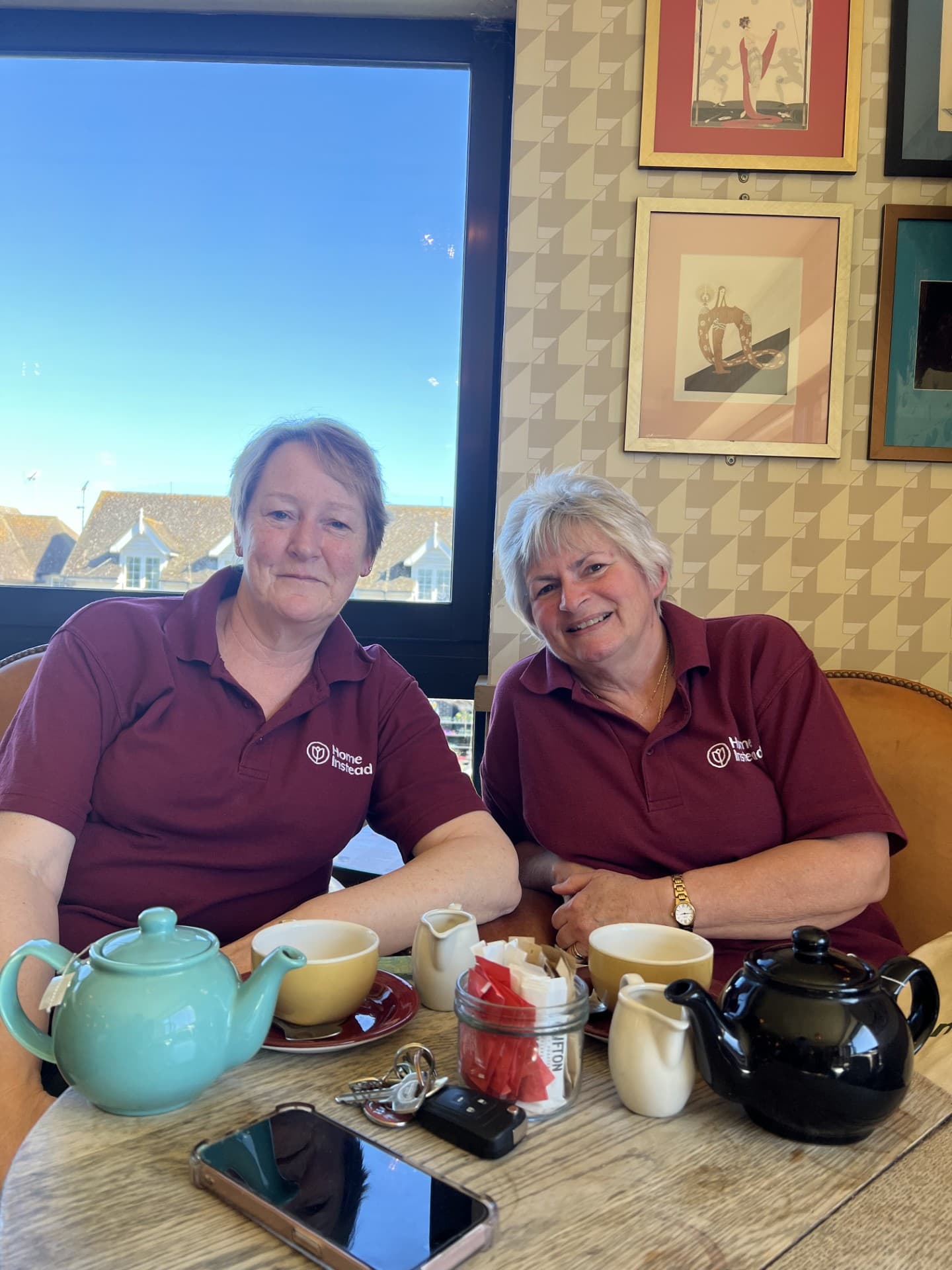 Two women in matching maroon shirts sit at a table with tea pots, cups, and sugar, smiling at the camera. - Home Instead