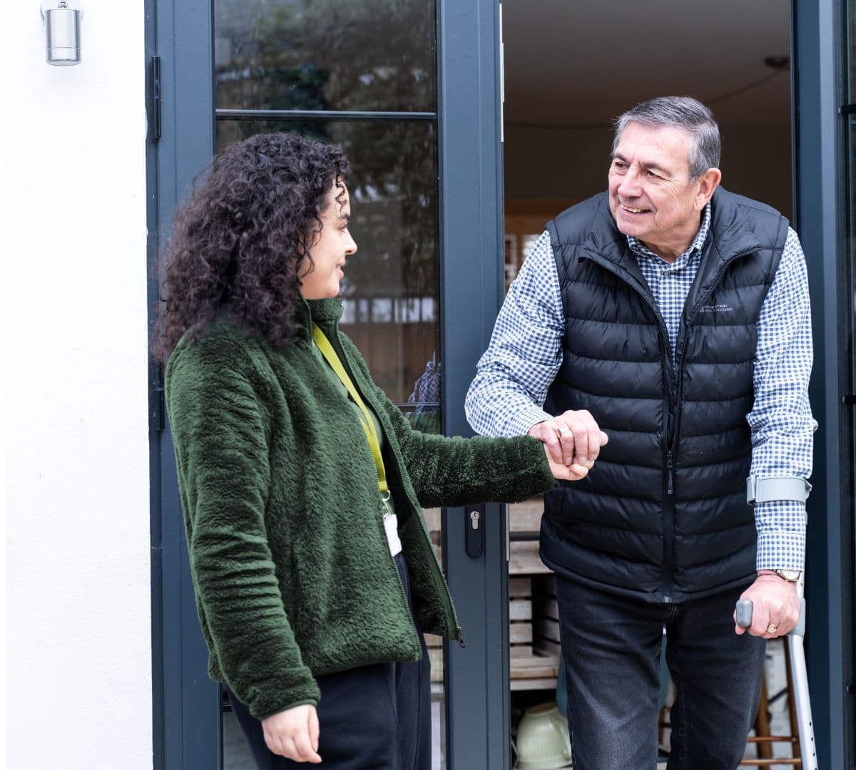 An older male adult with grey hair and wearing a puffer jacket adn using a crutch while walking at the door and being helped by his younger female carer wearing green and with black curly hair both happy and smiling