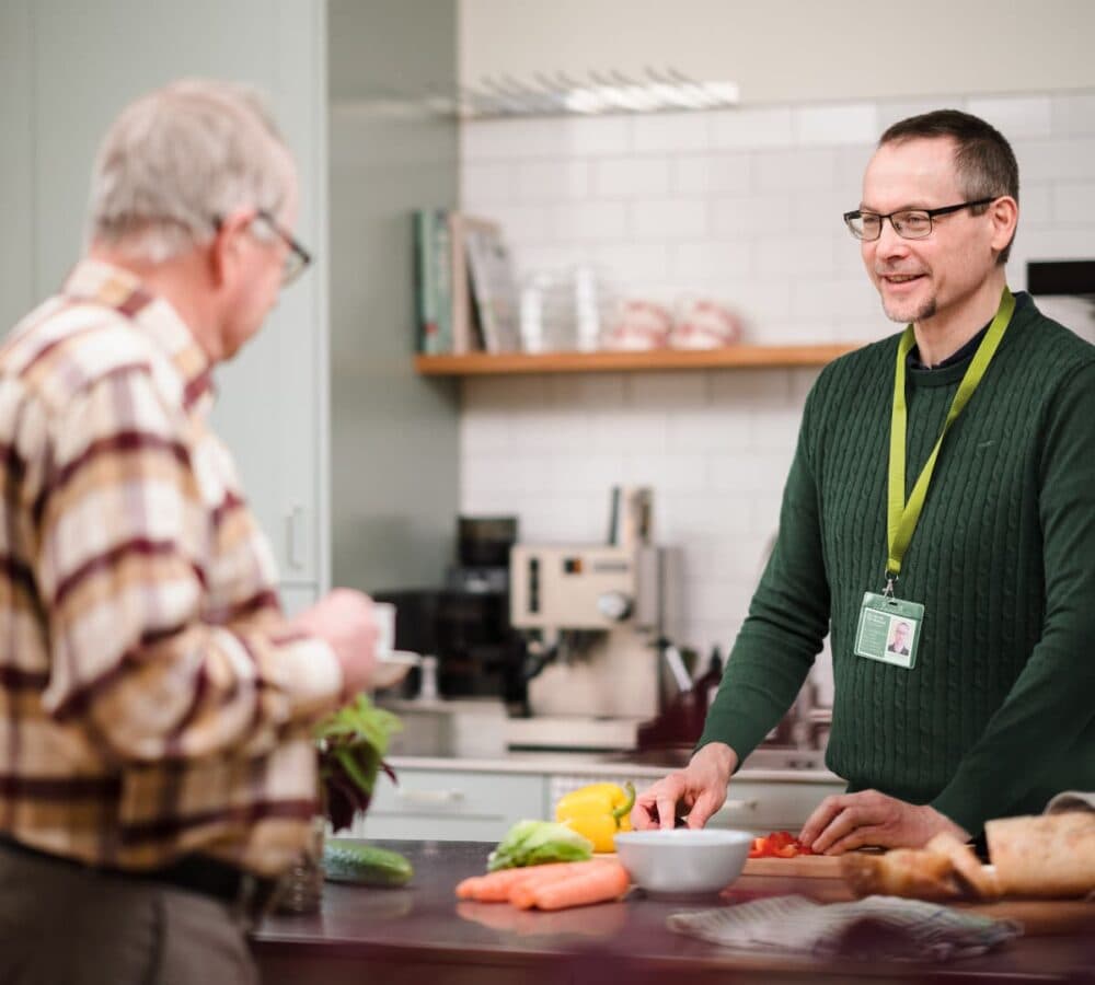An older male adult wearing brown and with eyeglasses inside the kitchen with his younger male carer with eyeglasses and wearing green while chopping some vegetables