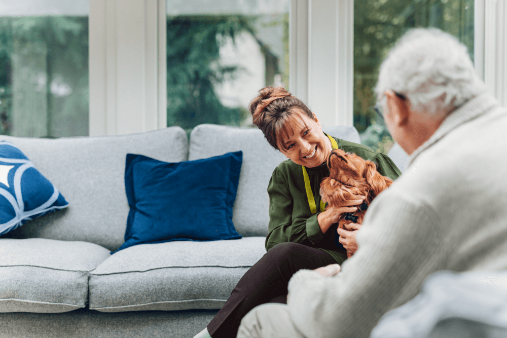 Elderly client and Care pro in the living room talking, there is a dog in the care pros lap. Home Instead Taunton and West Somerset Home care and Live in care