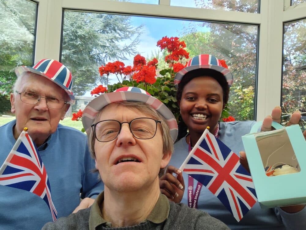 Three people in union jack hats holding British flags, smiling indoors with red flowers in the background. - Home Instead