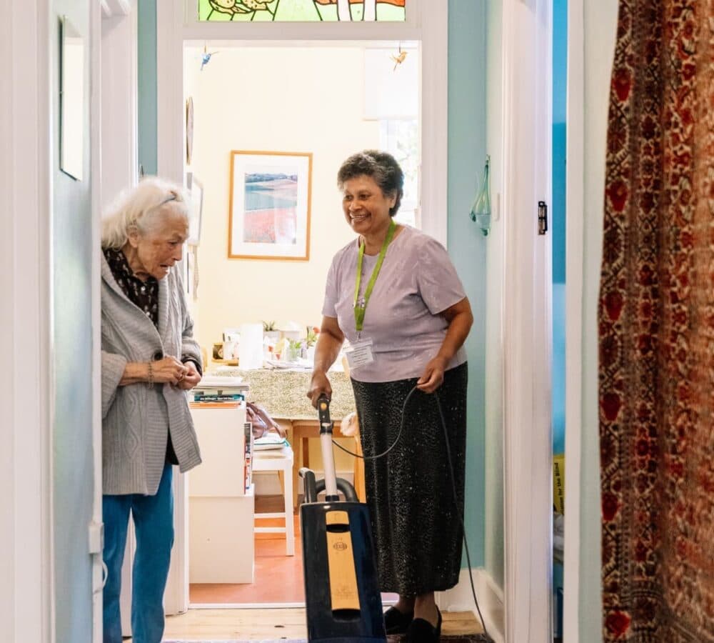A woman with short hair and wearing skirt cleaning and vacuuming the floor with an older female adult