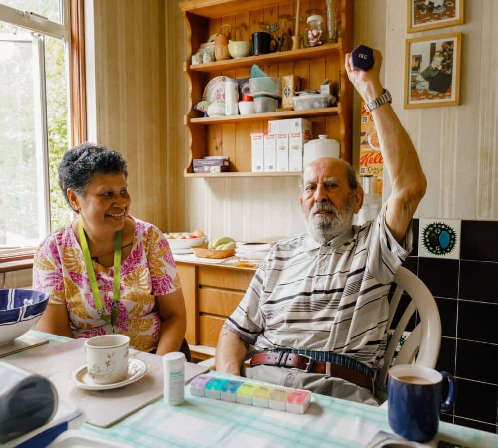 An older male adult exercising while sitting down in the kitchen with his younger female carer with black hair