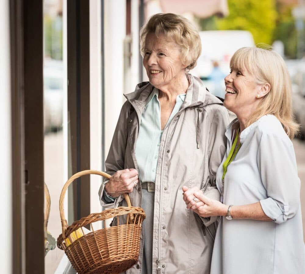 An older female adult with short hair, bringing a basket and wearing sweater with her younger female carer with blonde hair happy and smiling outdoors