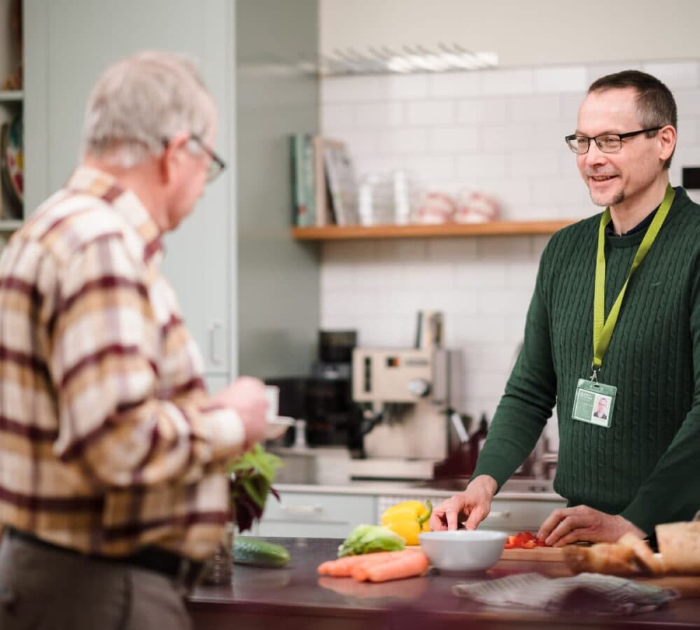 An older female adult holding a cup and wearing eyeglasses with grey hair chatting with his male carer wearing green and chopping vegetables inside the kitchen