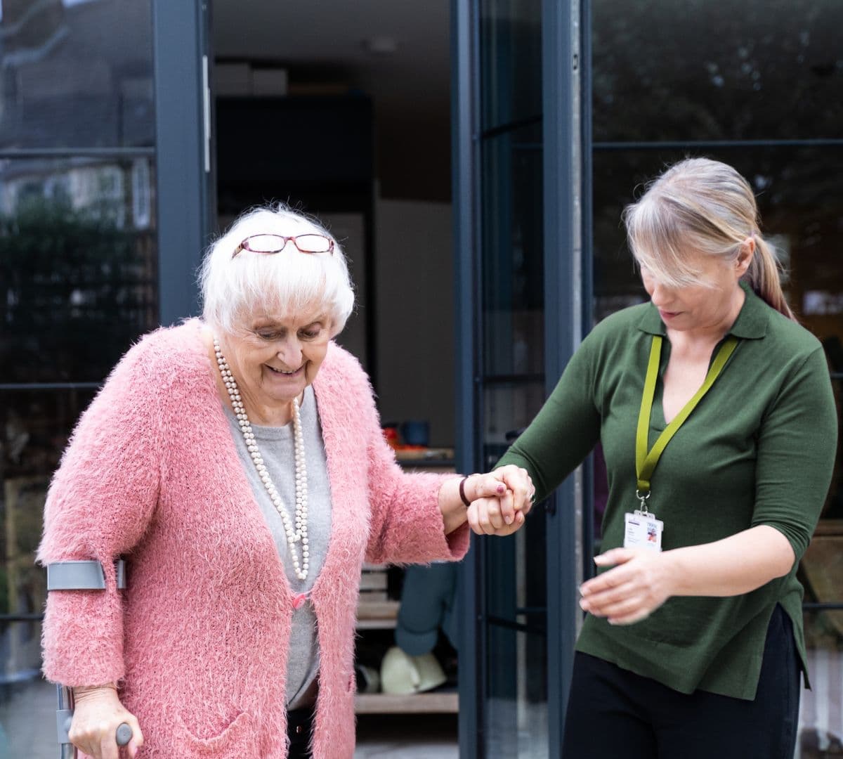 An older female adult with white hair and wearing a pink sweater while holding a crutch to walk and being assisted by her younger female carer wearing green