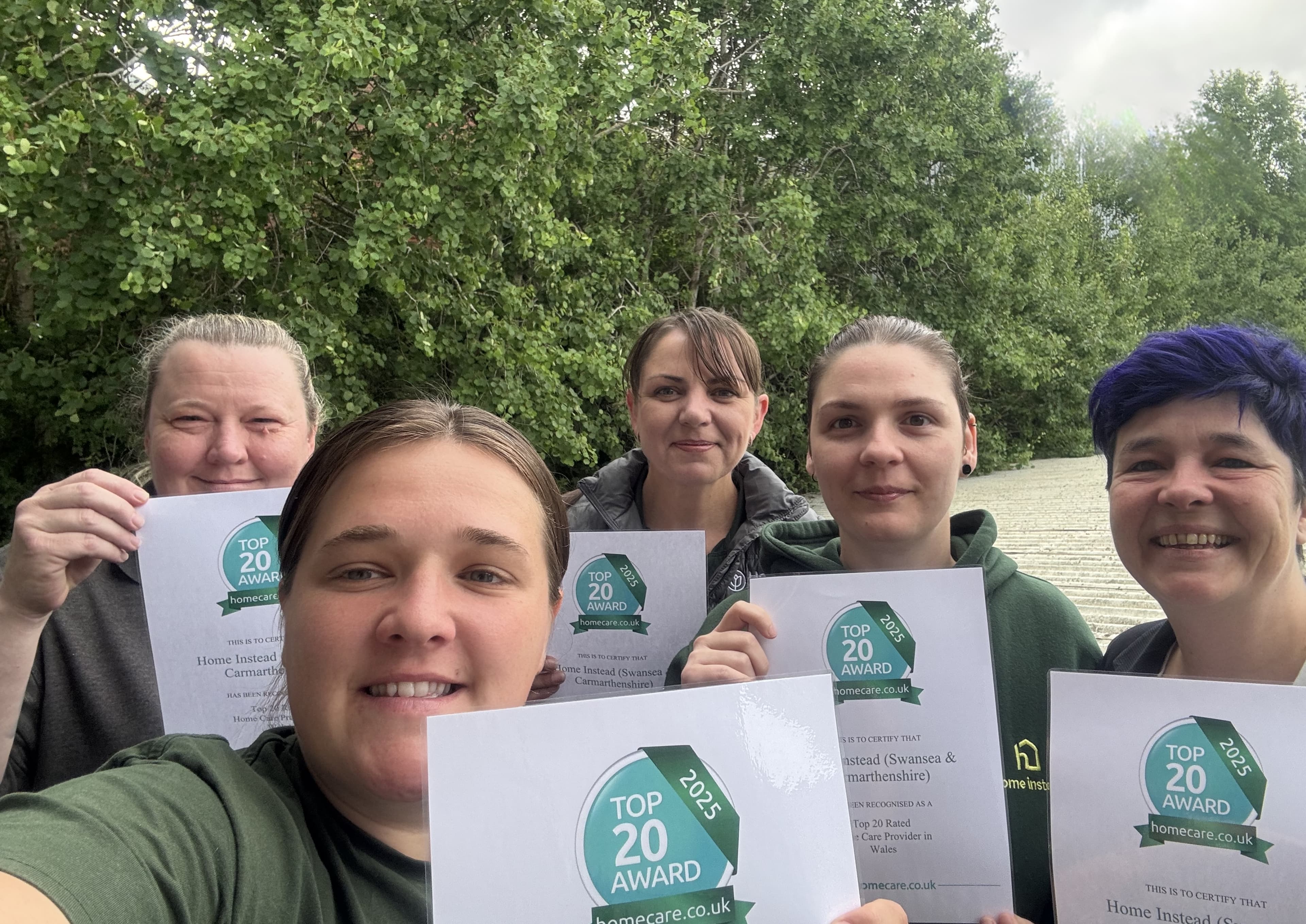 Group of people holding certificates for the "Top 20 Award 2023" from homecare.co.uk, standing outdoors in front of a lush green background.
