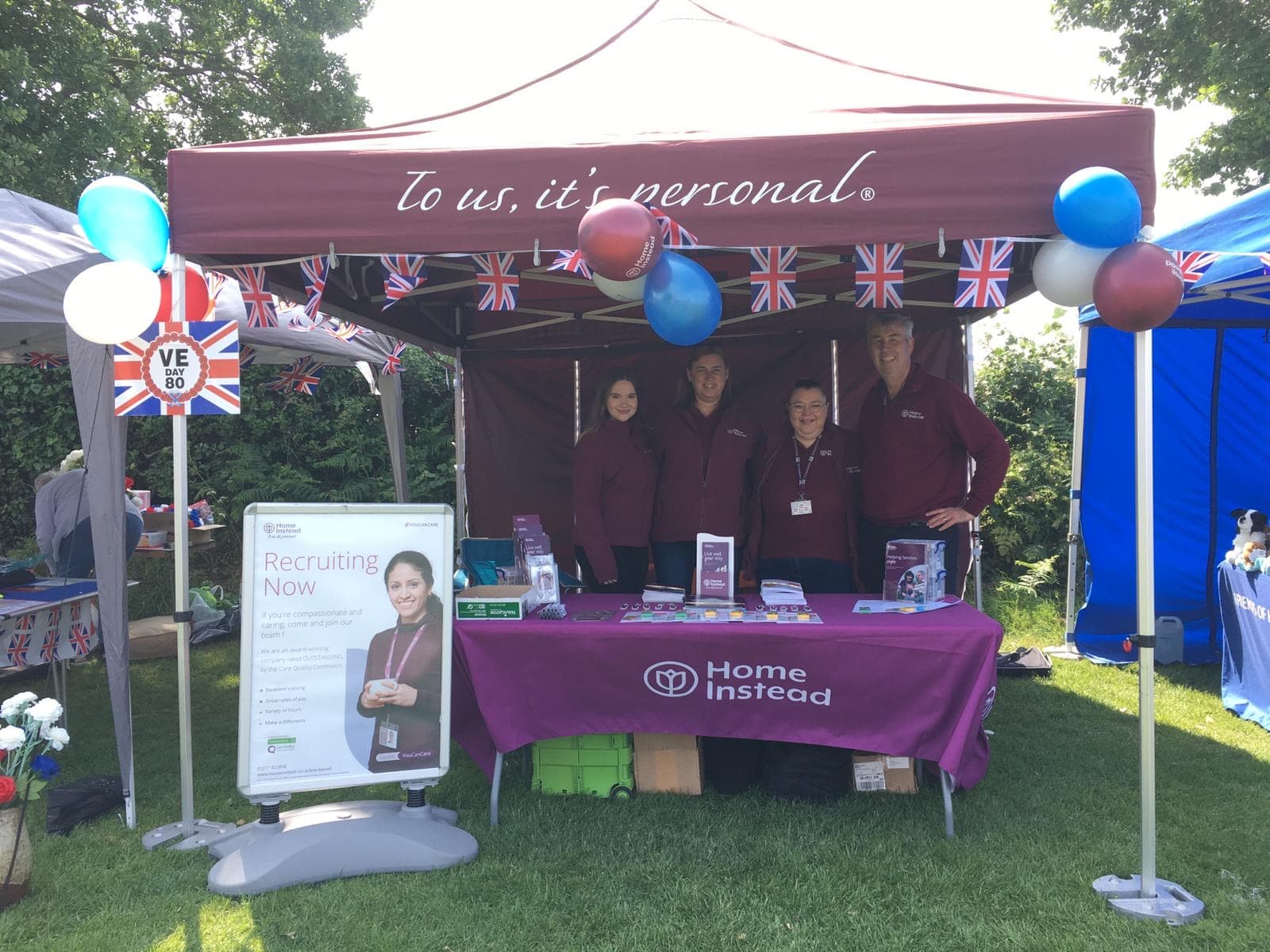 Four people stand at a Home Instead recruitment booth outdoors, decorated with balloons and bunting. - Home Instead