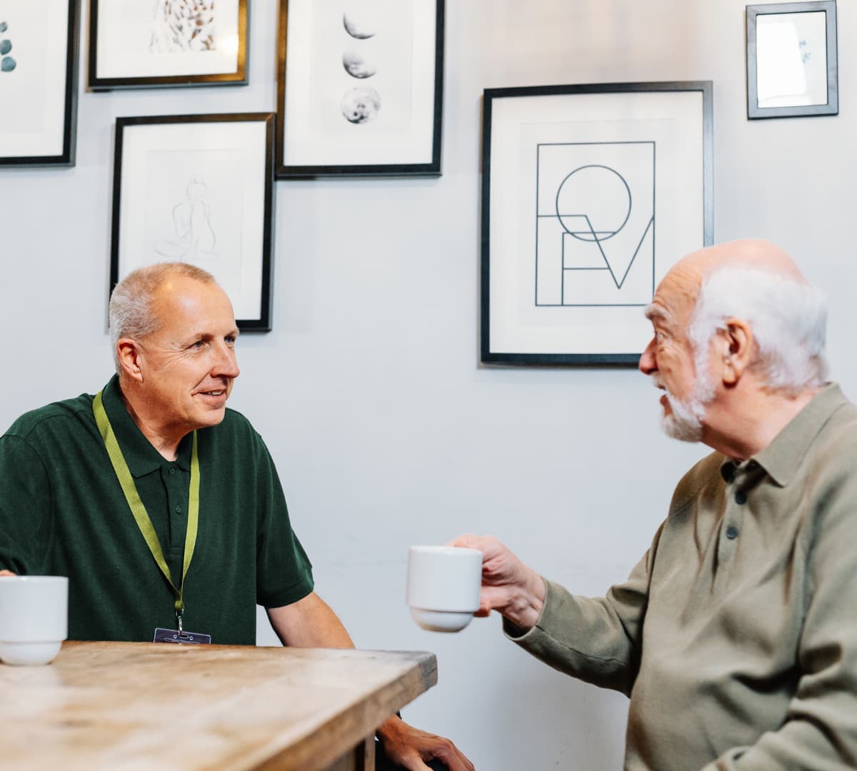 An older male adult with grey hair drinking coffee with a younger female carer wearing green both chatting with each other