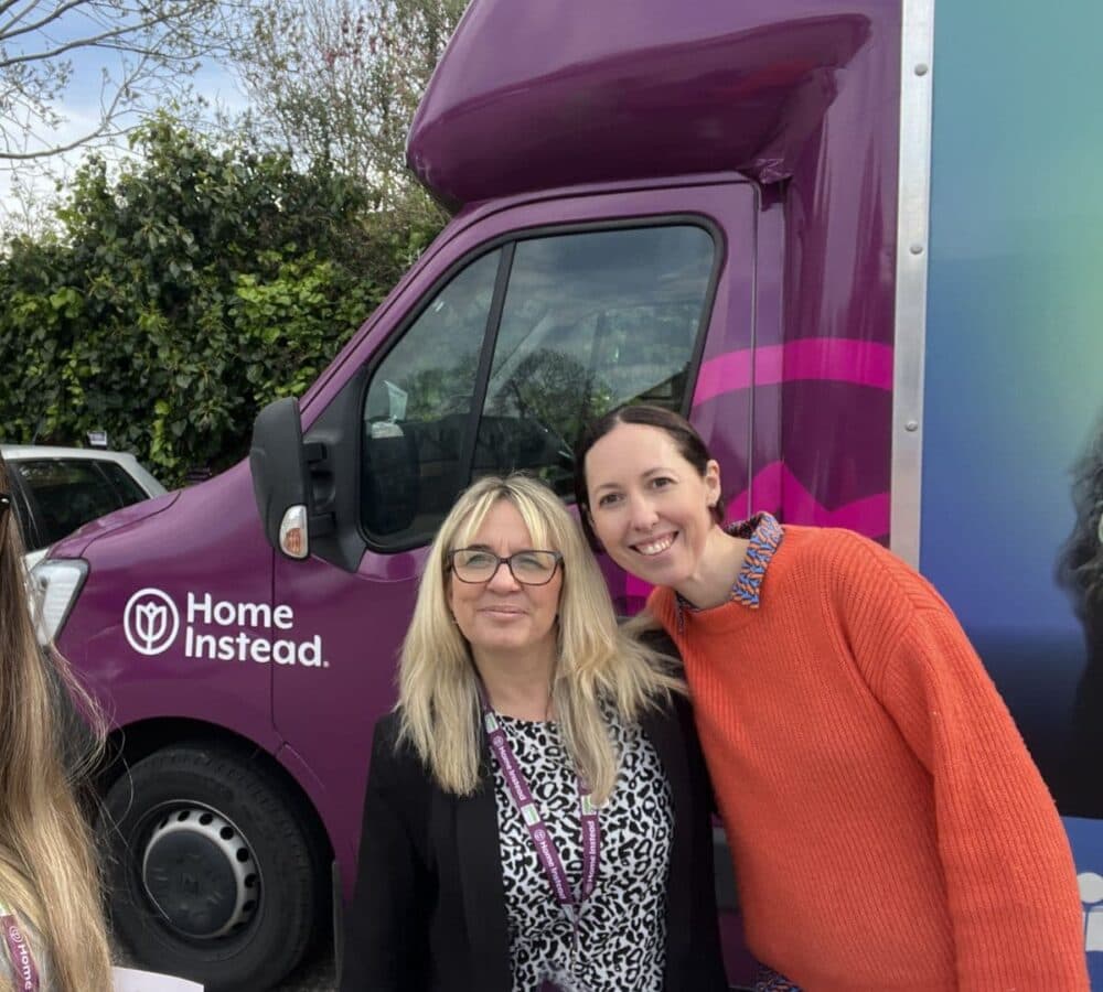 Two women both smiling with a purple bus at their back