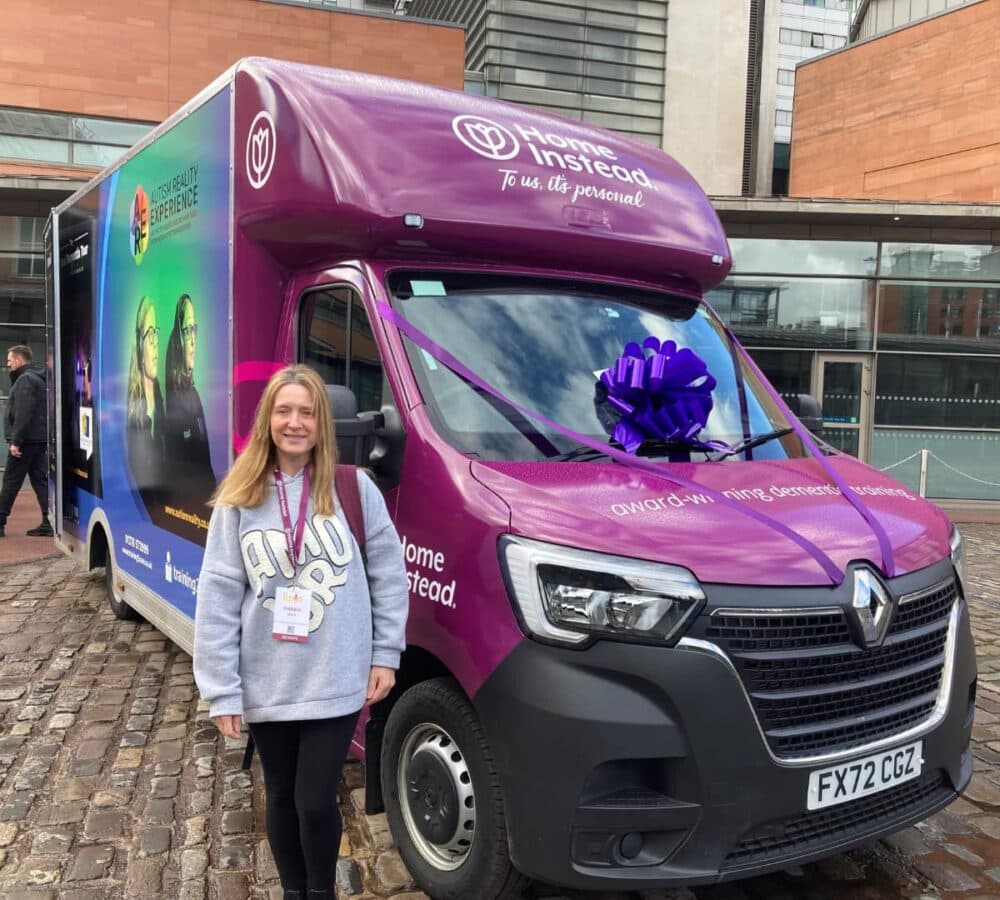 Woman with long hair wearing pullover jacket smiling with a purple bus beside her