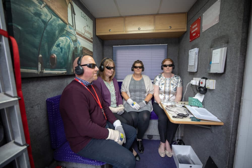 Four people sit in a room wearing dark glasses and headphones, each holding a white cane and wearing gloves. - Home Instead