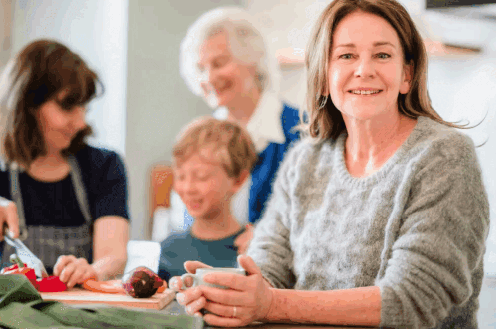 Smiling woman sits at a table, with family members preparing food together in the background. - Home Instead