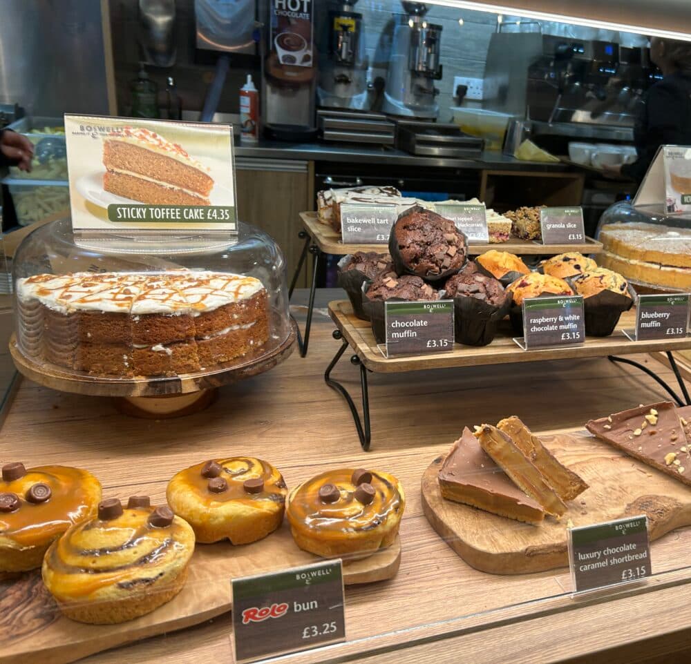Cakes counter at Boswells cafe in Basingstoke showing sticky toffee cake, muffins of various flavours, Rolo buns, chocolate caramel shortbread and a selection of other cakes & pastries