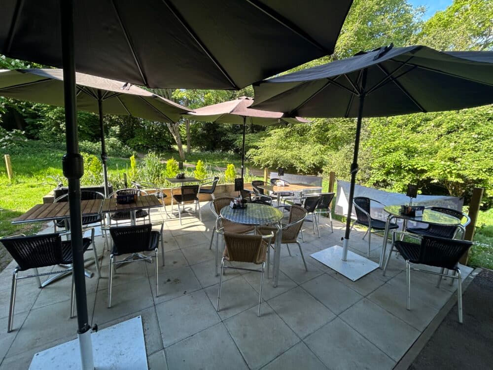 An outdoor seating area showing tables and chairs under four large umbrellas on a tiled floor, with parkland in the background