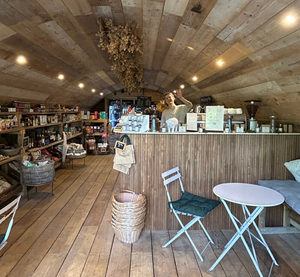 Interior view looking down the length of the Trough Farm Shop, showing a timber arch ceiling, timber floor, goods on the shelves and a man waving from behind the counter