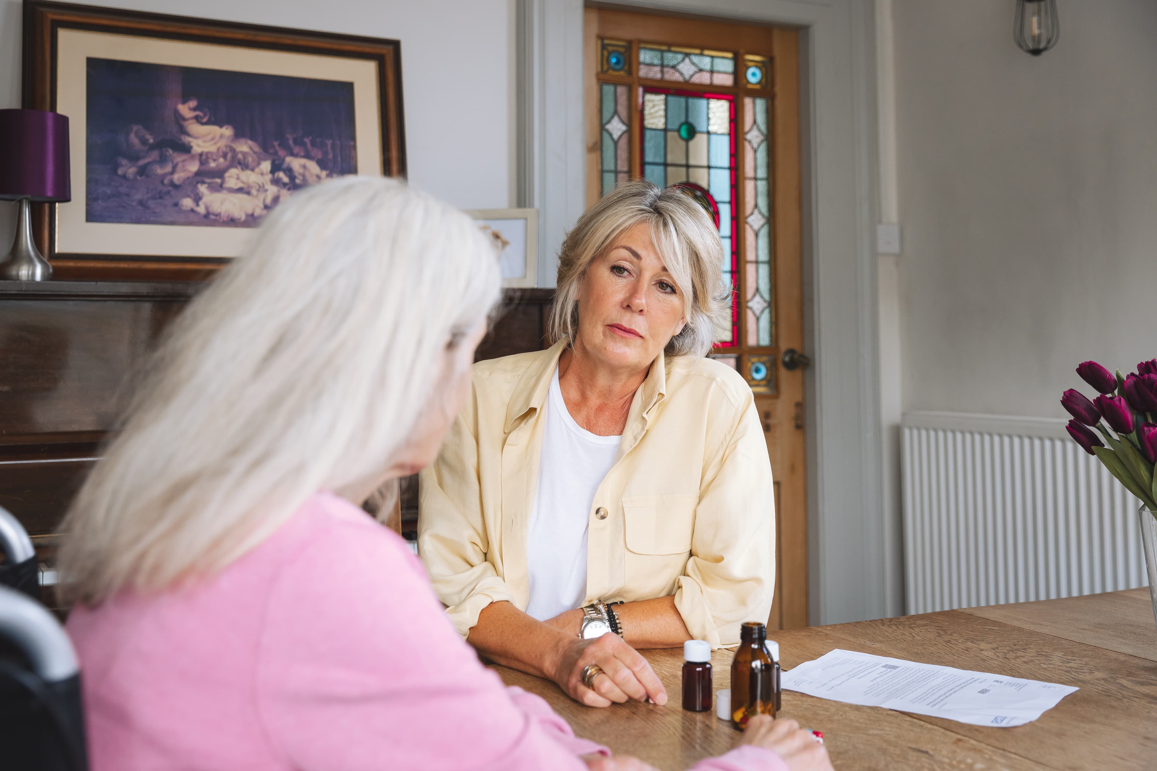 Two women sit at a table, talking seriously, with medicine bottles and papers in front of them. - Home Instead