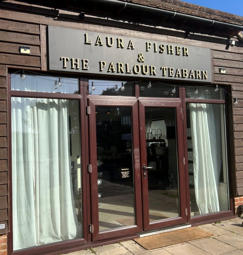 Large glass doors set in the end of a wooden barn with a name board above the door saying "Laura Fisher and The Parlour Teabarn", with pictures of homeware at each side of the doors.