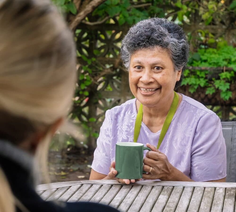 A woman smiling while drinking a coffee using a green mug outside the garden