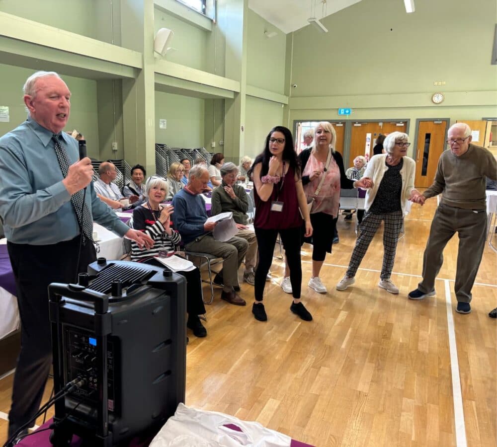 A man sings into a microphone while seniors and others dance and watch in a community hall. - Home Instead