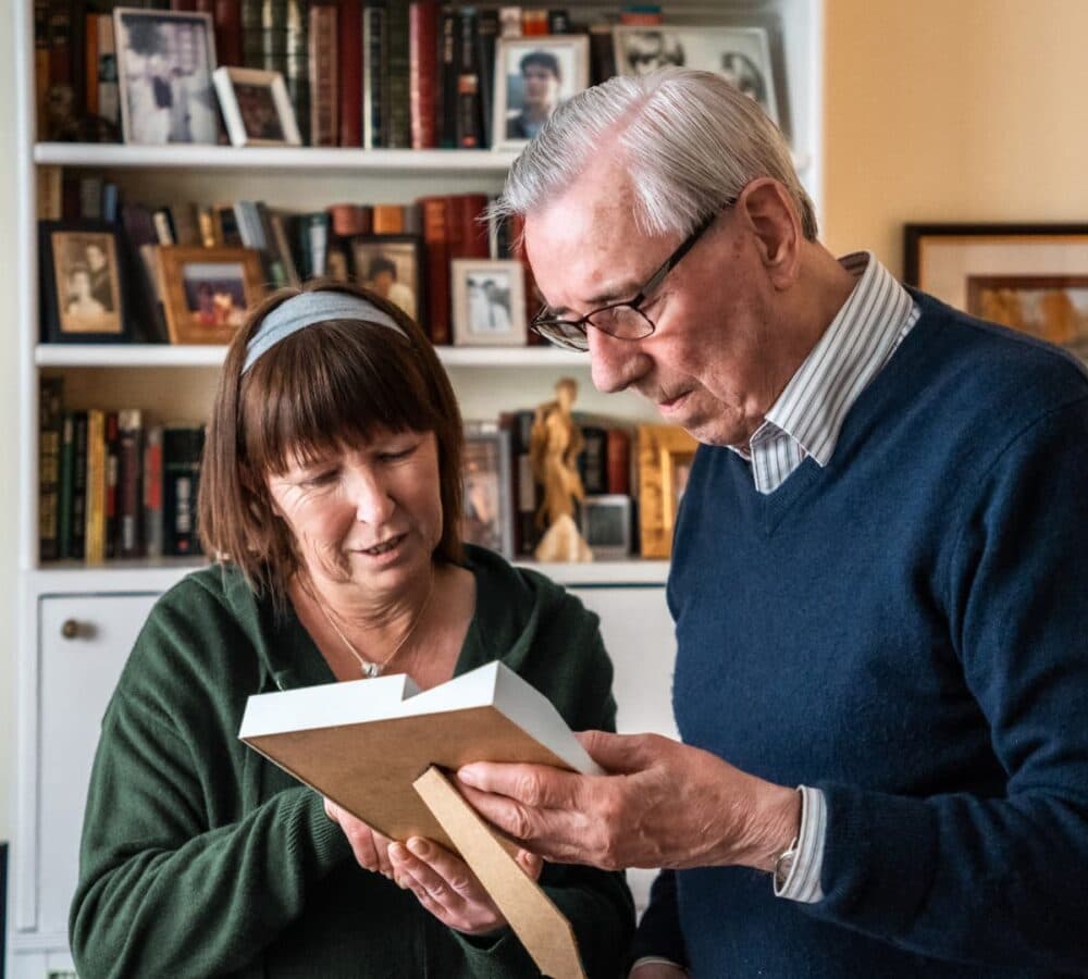 An older male adult wearing eyeglasses with his young female carer with short hair both reading a book