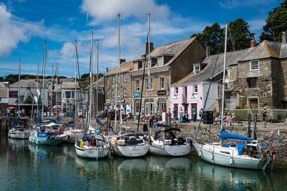 Boats docked at a harbor with stone buildings and shops along the waterfront under a blue sky. - Home Instead
