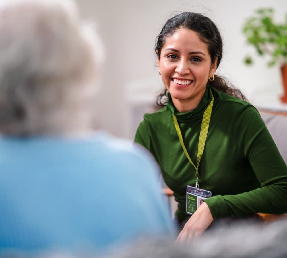 A woman with long curly hair wearing green happy and smiling while sitting on a couch inside the house